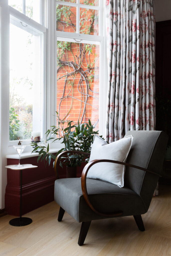 A modern gray chair with wooden arms sits beside a small table holding a martini glass. Nearby, a potted plant thrives in the sunny corner by large windows framed by floral curtains.