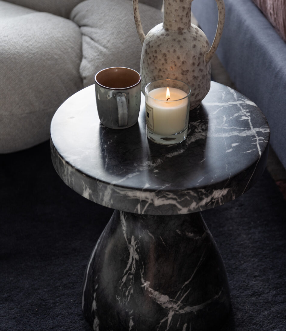 A round black marble table holds a decorative pitcher, a ceramic mug, and a lit candle, set on a dark rug beside a light-colored, plush armchair.