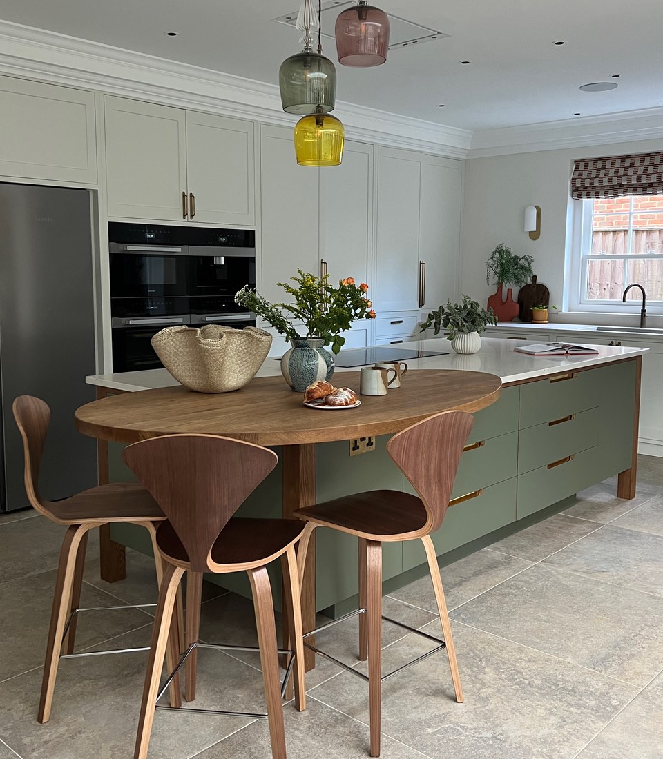 A round wooden table with two modern chairs holds a vase of flowers and a plate of pastries. The kitchen features light cabinets, stylish pendant lights, and a bright window.