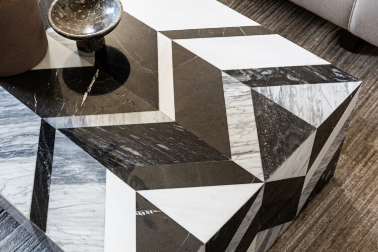 A geometric marble coffee table, featuring alternating black and white triangular patterns, sits on a textured beige rug in front of a light-colored sofa. A small bowl and a vase with greenery are placed on top.