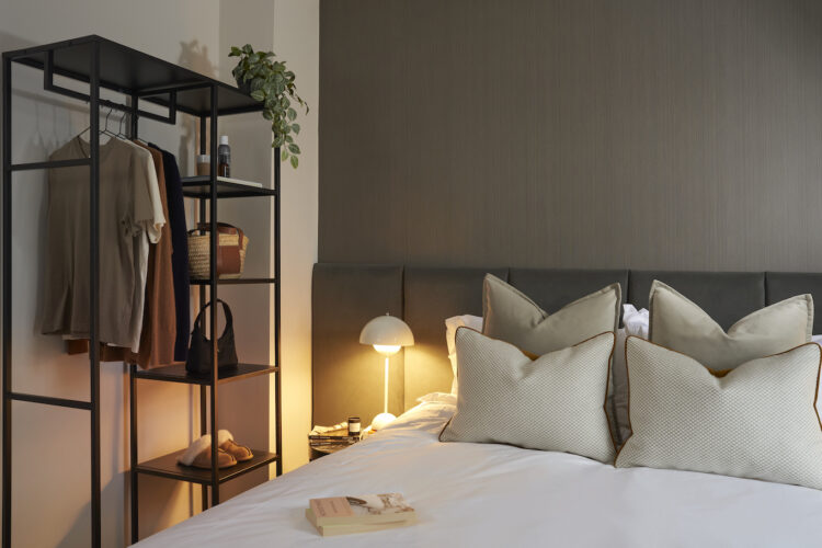 A stylish bedroom features a black metal shelving unit with clothes and accessories, a cozy bed with plush pillows, and a soft bedside lamp casting warm light, enhancing a serene atmosphere.