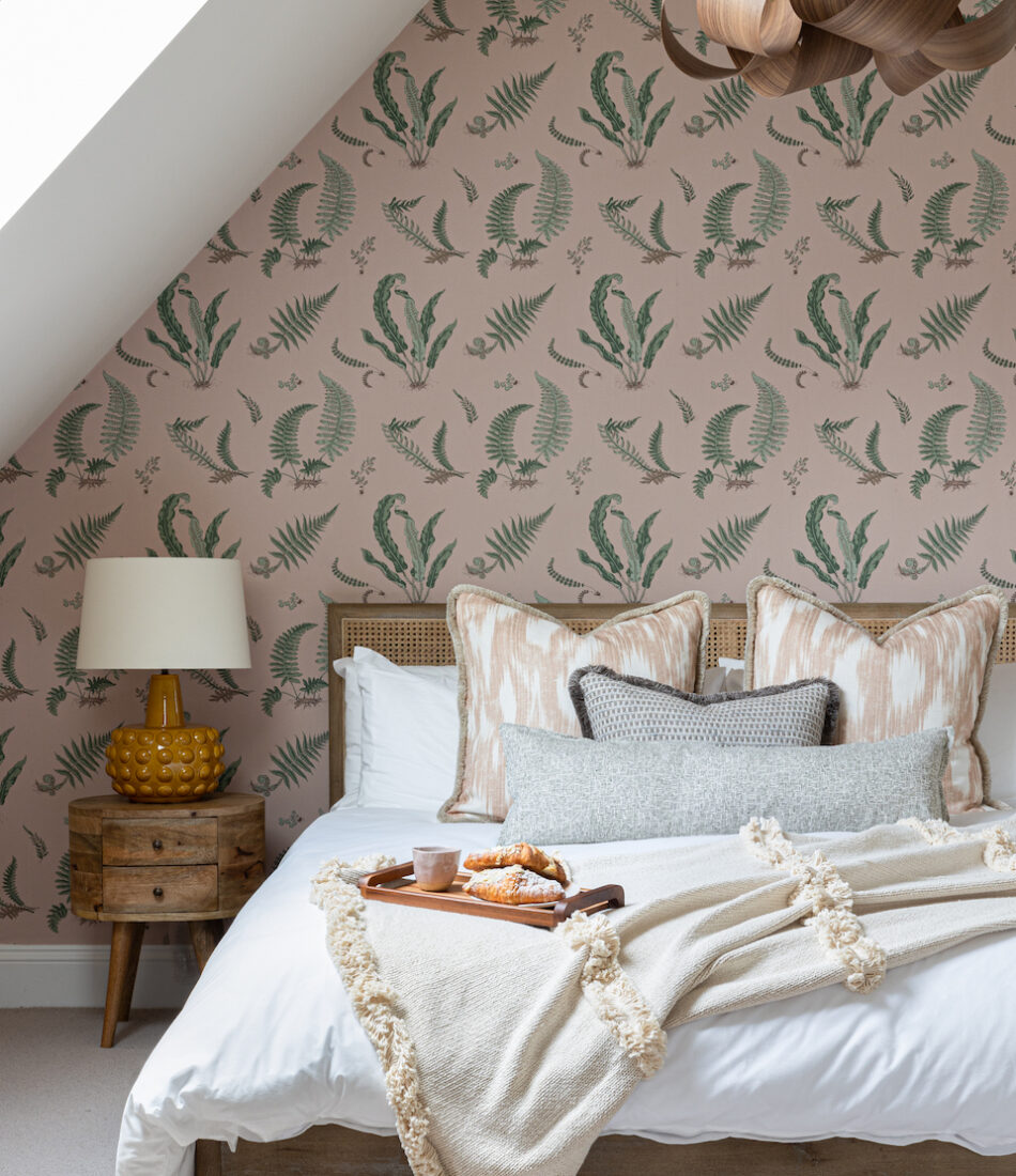 A cosy bedroom features a bed with decorative pillows and a blanket, a wooden bedside table with a lamp, and a tray of pastries. The walls are adorned with botanical-patterned wallpaper.