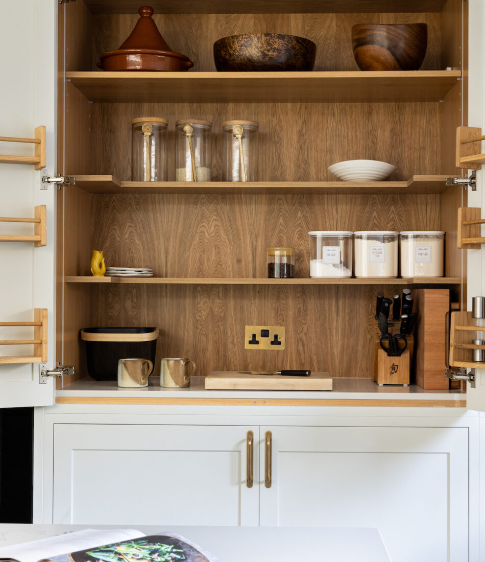 A kitchen cabinet displays various items including a terracotta pot, wooden bowls, glass jars, and coffee containers. A countertop holds a cookbook and cutting board, with utensils neatly arranged.