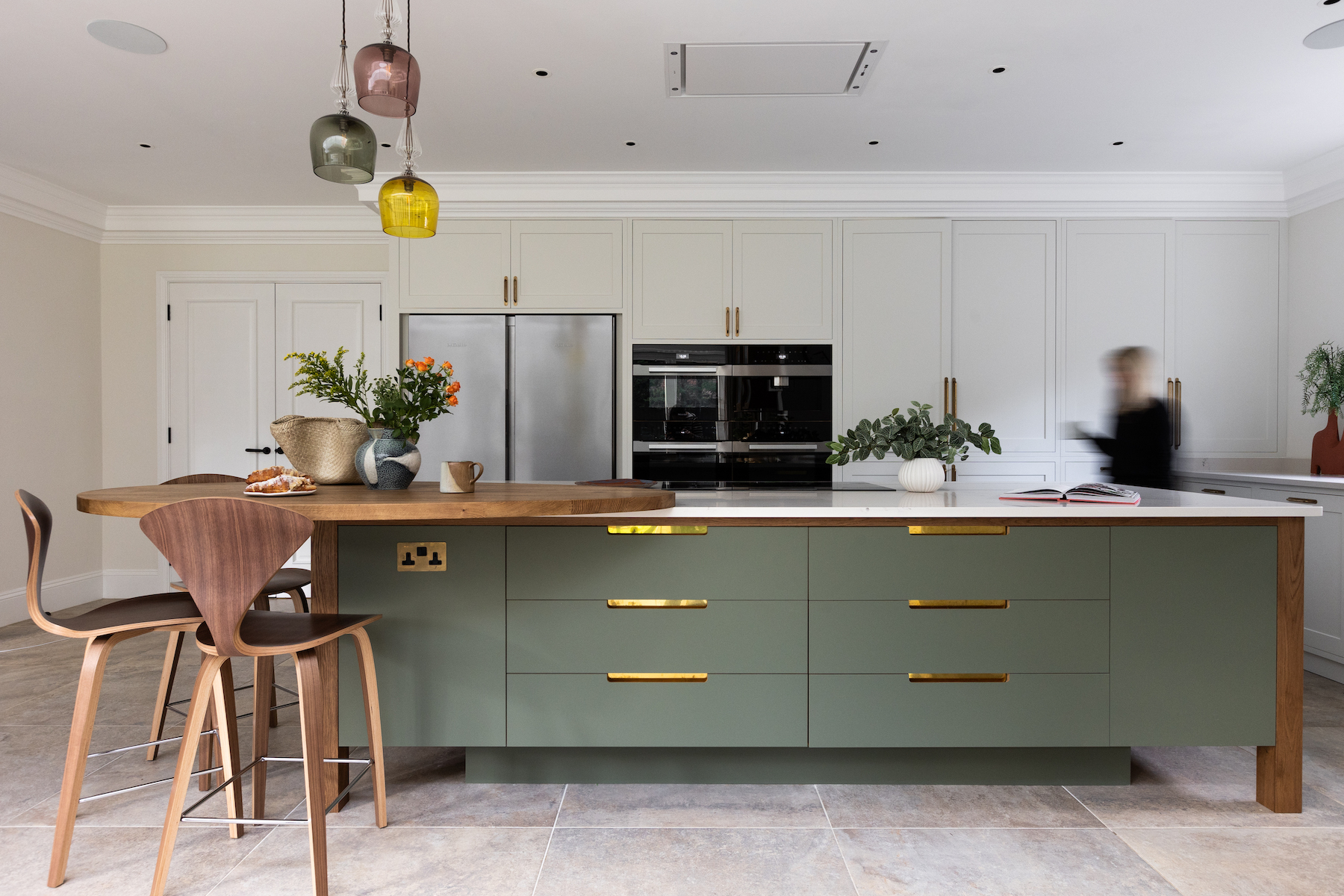 A green kitchen island with wooden countertops displays a vase of flowers and baked goods. A person in motion passes by, while colorful pendant lights hang above. The context is a modern kitchen setting.