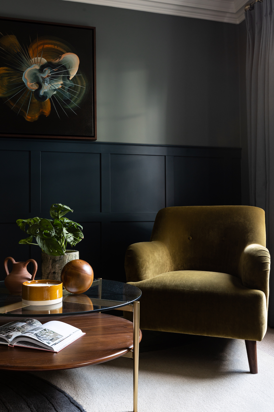 A green velvet armchair sits beside a round coffee table, displaying a candle and decorative items, in a cozy living room with dark paneled walls and a large abstract painting.