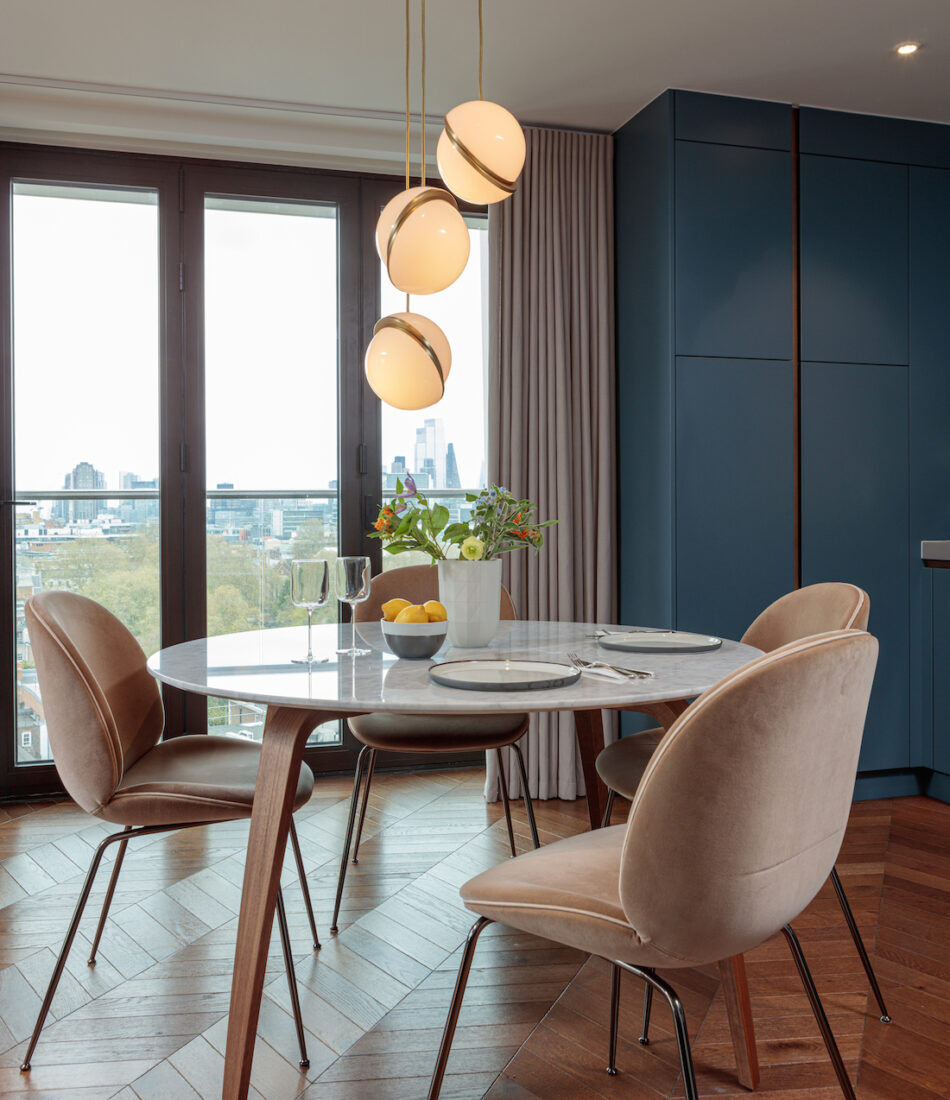 A round marble dining table with elegant chairs is set with glassware and plates. A bouquet of flowers and lemons adorn the table, illuminated by hanging pendant lights, against a scenic urban view through large windows.