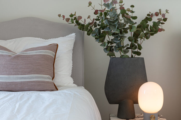 A plush bed with decorative pillows sits beside a marble side table. An abstract art piece adorns the wall, while a modern lamp and greenery complete the cozy bedroom scene.