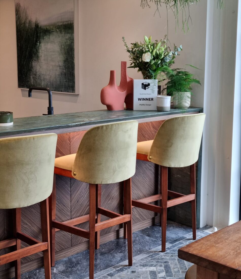 Three yellow-green bar stools are positioned at a wooden counter with a textured backdrop. A plant arrangement and a decorative vase are atop the counter, amidst natural lighting and a modern setting. A sign reads: "WINNER Pfeffer Design."
