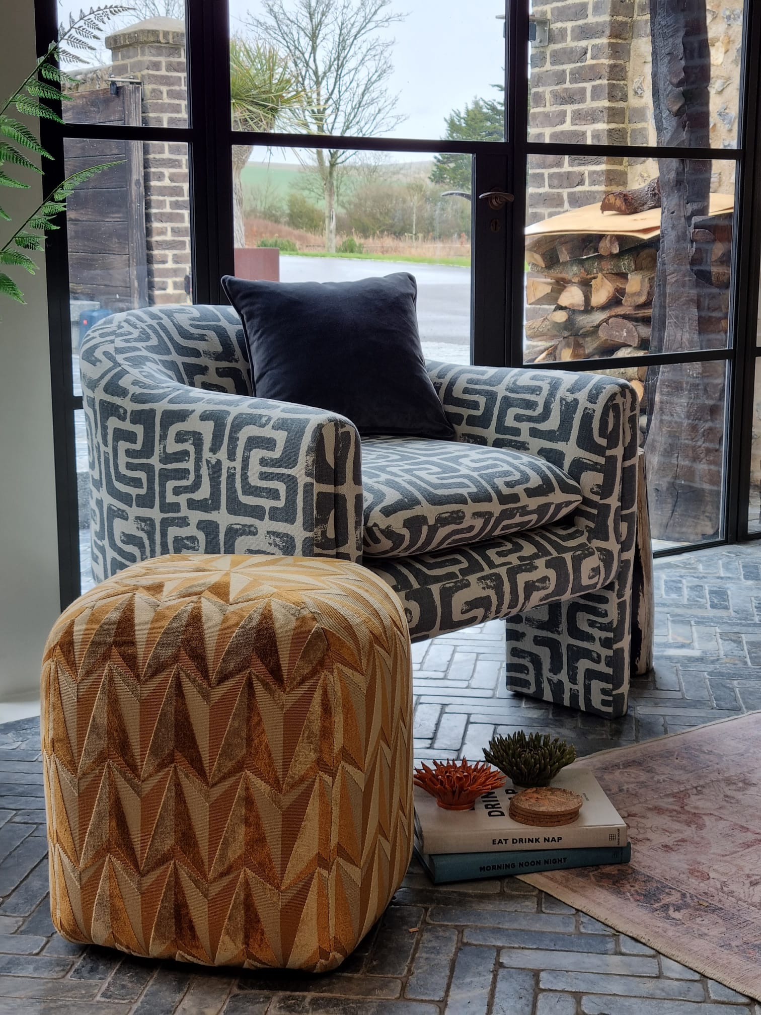 A patterned armchair with a dark cushion sits beside a geometric pouf on a textured floor, with books and decorative plants nearby, all framed by large windows showing an outdoor scene.