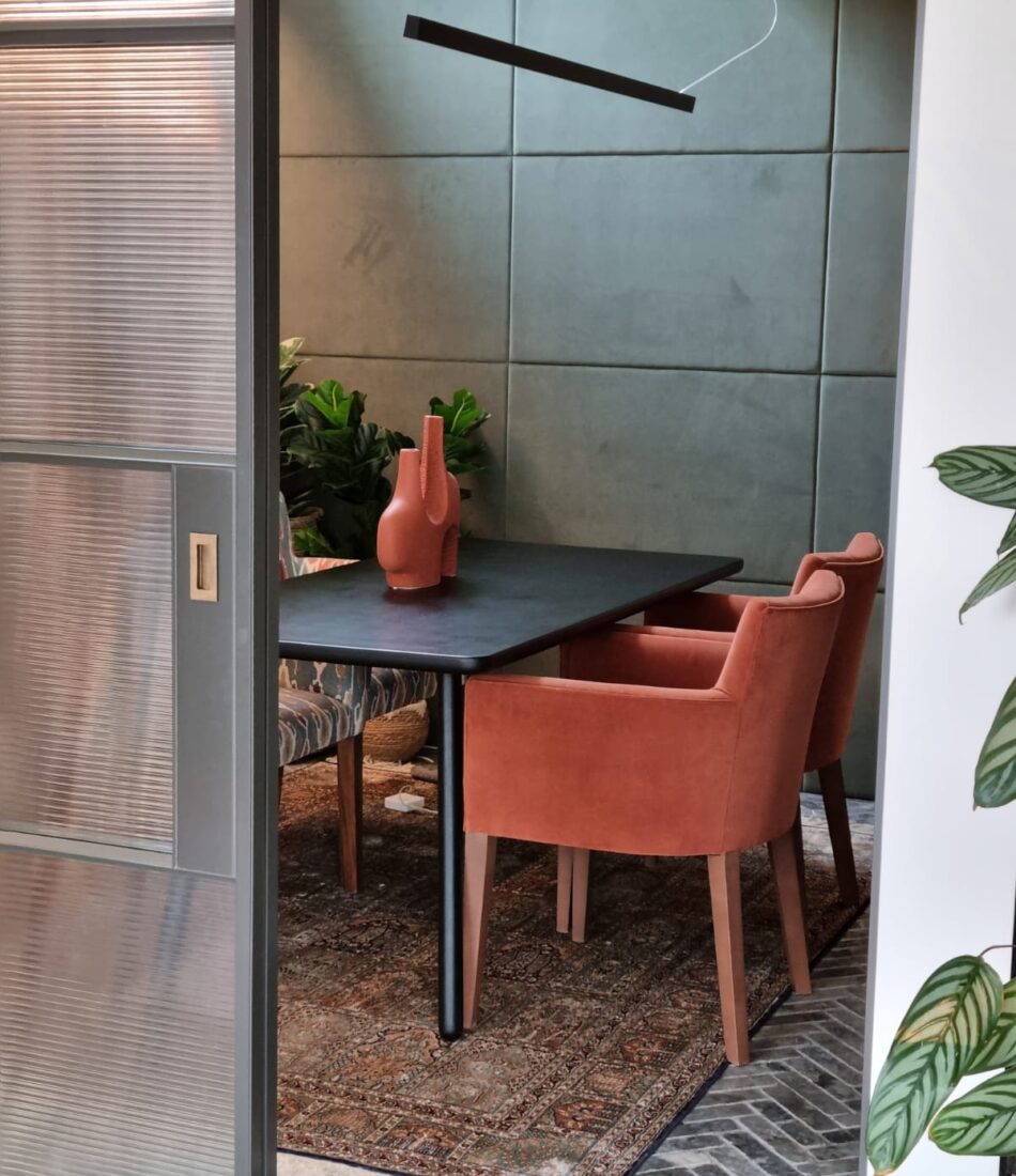 A black table with two coral armchairs sits on a patterned rug in a cozy dining area, surrounded by green plants and a lightly textured blue wall, illuminated by a sleek overhead light.
