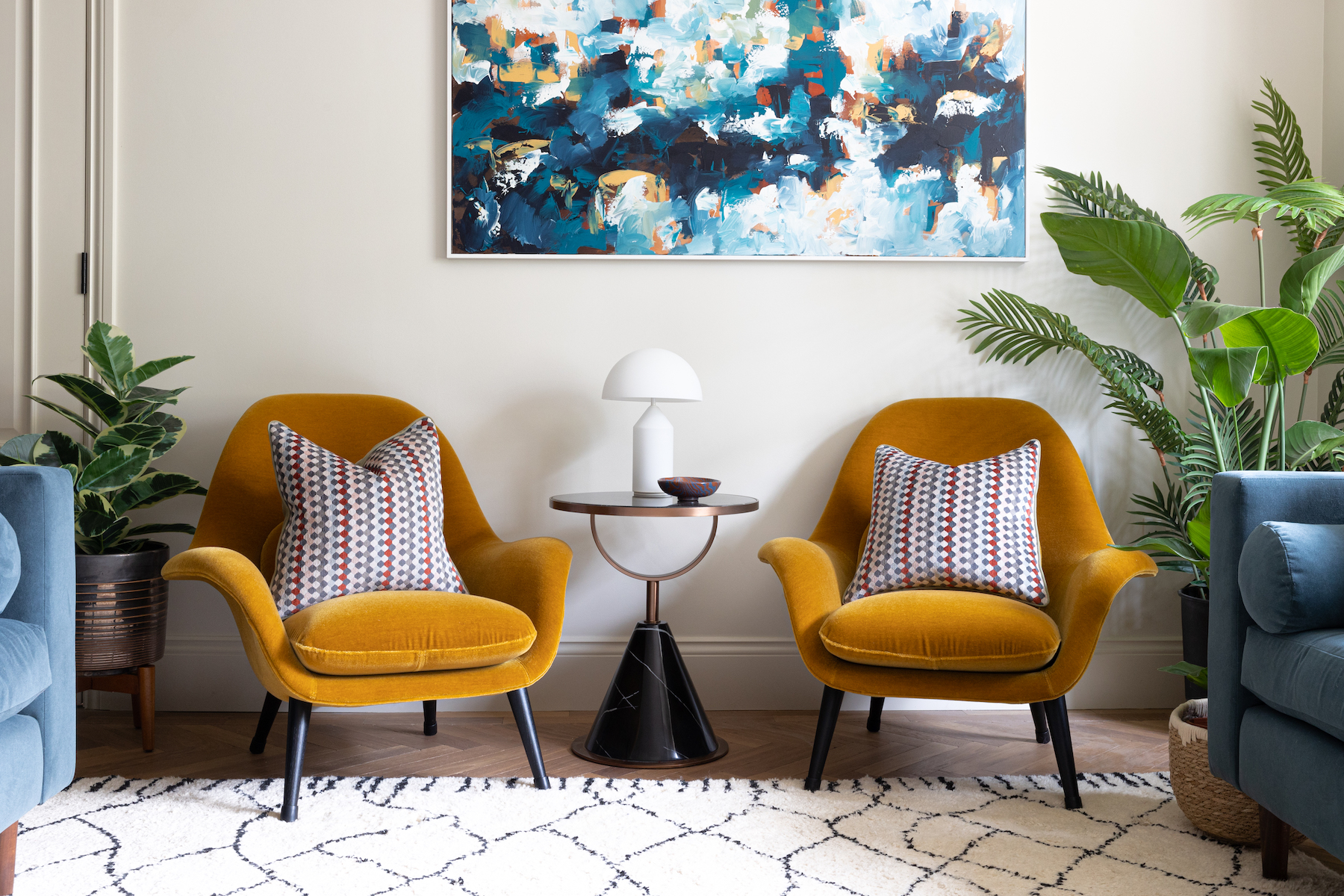 Two mustard yellow armchairs with patterned pillows face a small black side table topped with a white lamp, surrounded by potted plants, in a cozy, well-decorated living room. A colorful abstract painting hangs above.