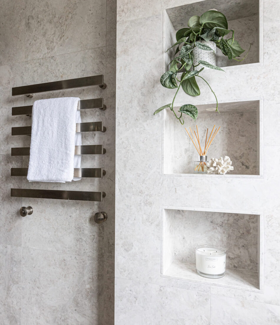A sleek bathroom wall features a towel rack holding a white towel and showcases stylish niches with a plant, decorative items, and a candle, all set against a light stone backdrop.