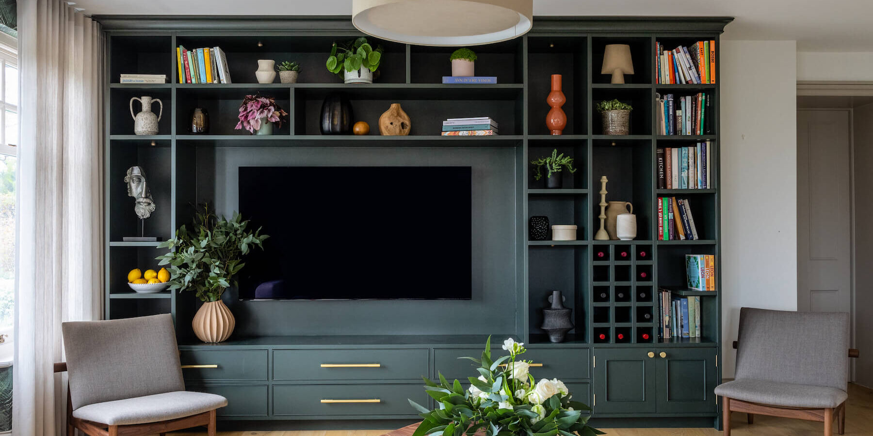 A green bookshelf with decorative items and a television is mounted centrally. Two armchairs face a wooden coffee table adorned with a floral arrangement and magazines in a bright room.