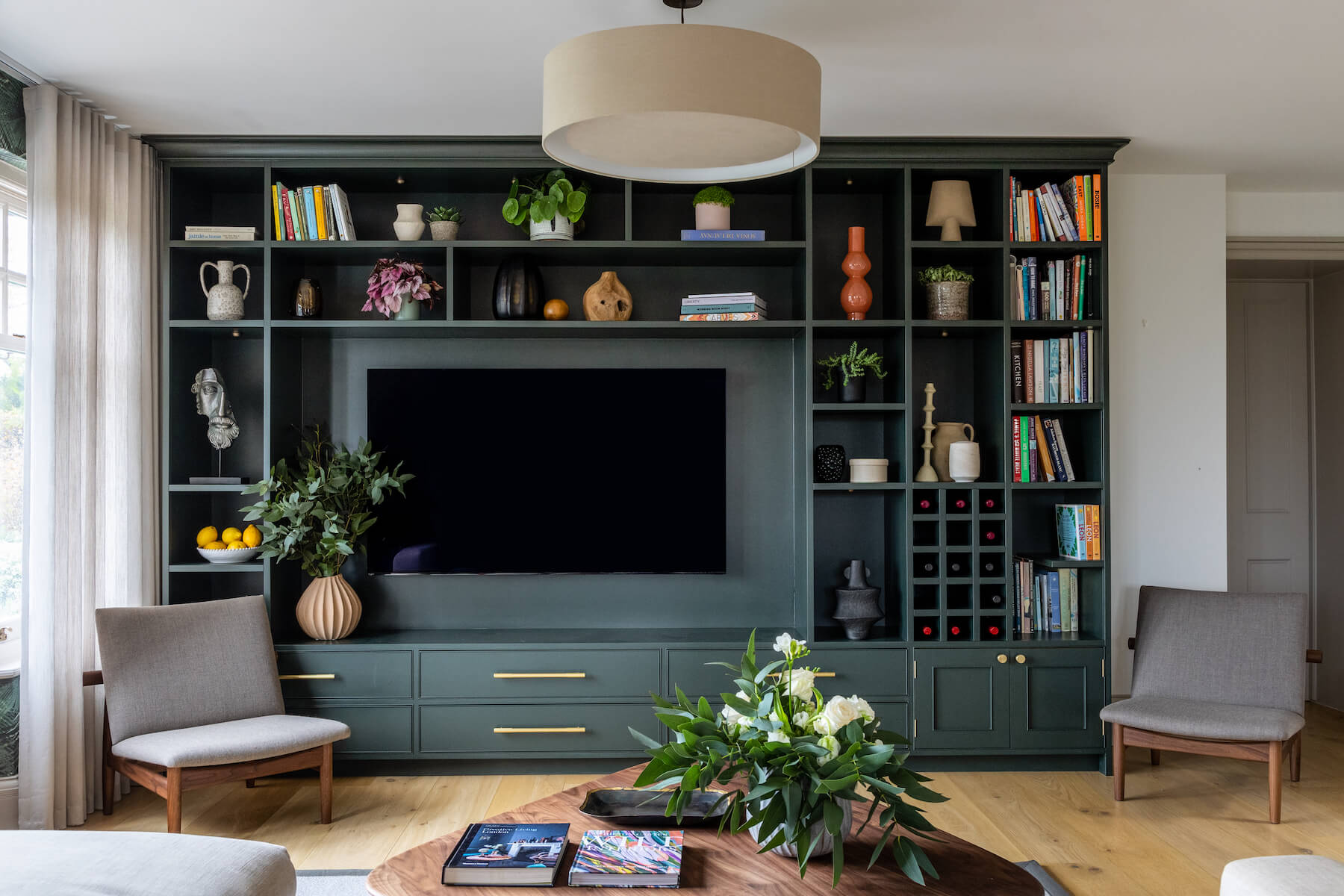 A green bookshelf with decorative items and a television is mounted centrally. Two armchairs face a wooden coffee table adorned with a floral arrangement and magazines in a bright room.