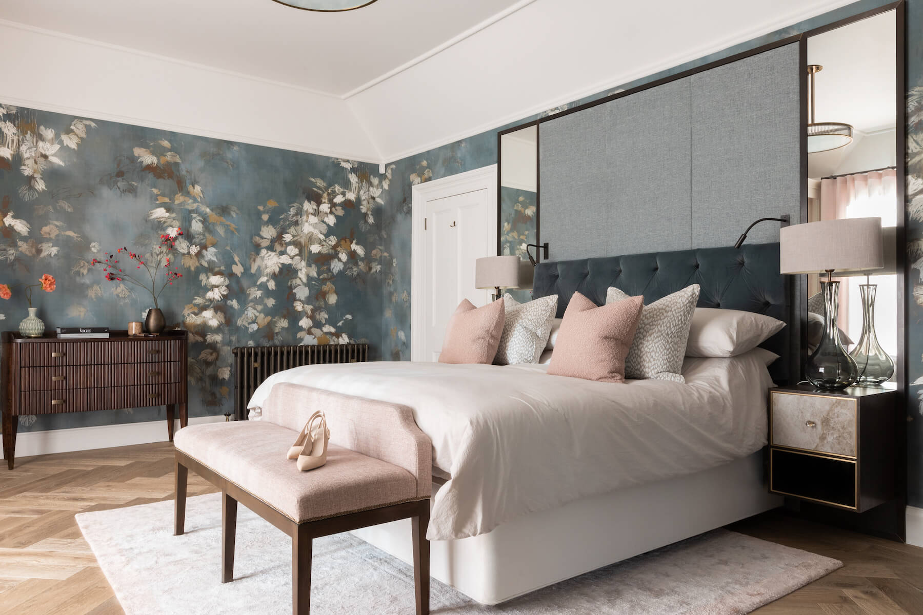 A stylish bedroom features a large bed with decorative pillows, a bench with a single shoe, and a floral wallpaper backdrop. Soft lighting illuminates a modern dresser and nightstand.