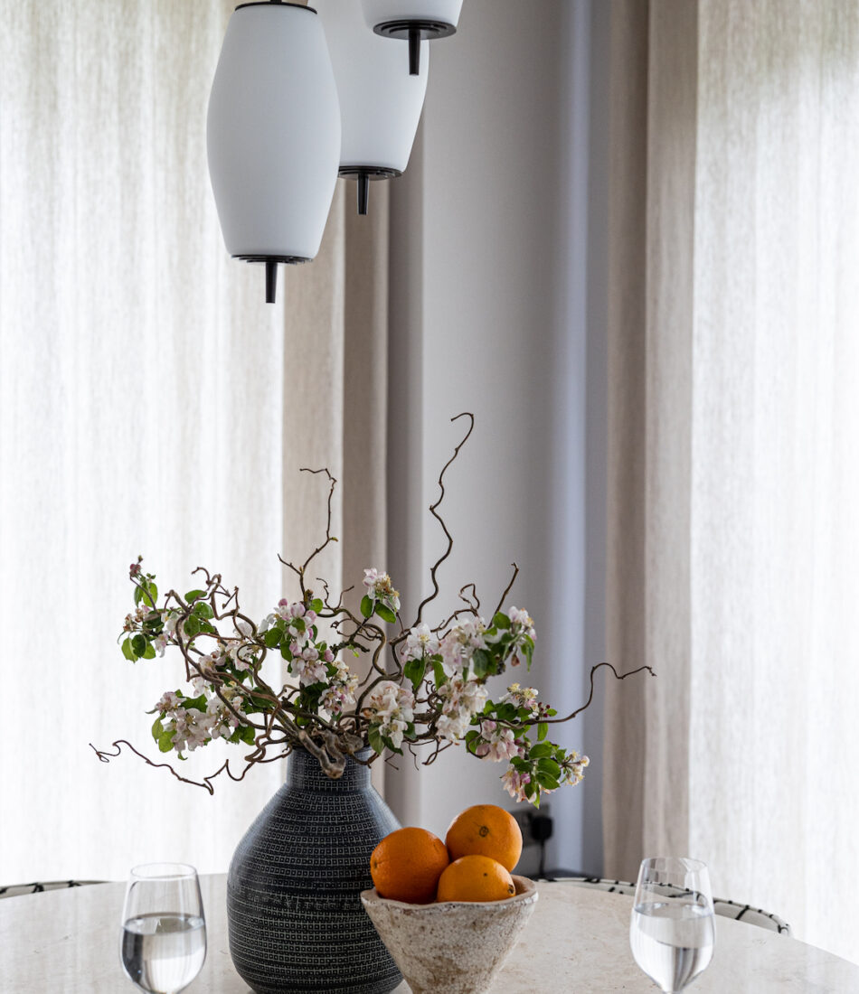 urban contemporary three glad drop pendant lights over a dining table with a bunch of cherry blossoms in a vase, a table setting for two and a bowl of oranges