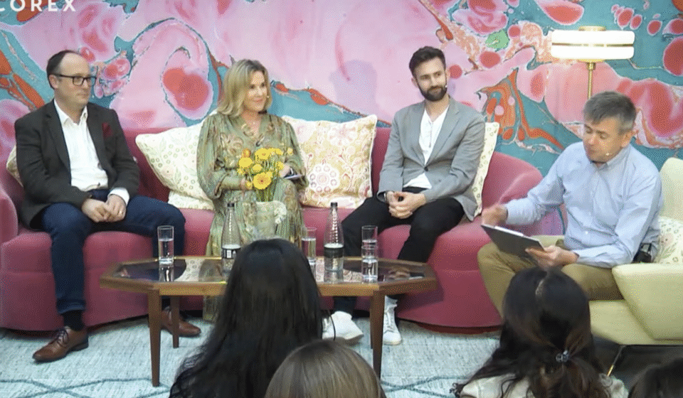 Four individuals are seated on a pink couch, engaged in discussion. One holds a notebook and flowers, while a table with glasses of water sits in front of them. A colorful abstract background sets the scene.