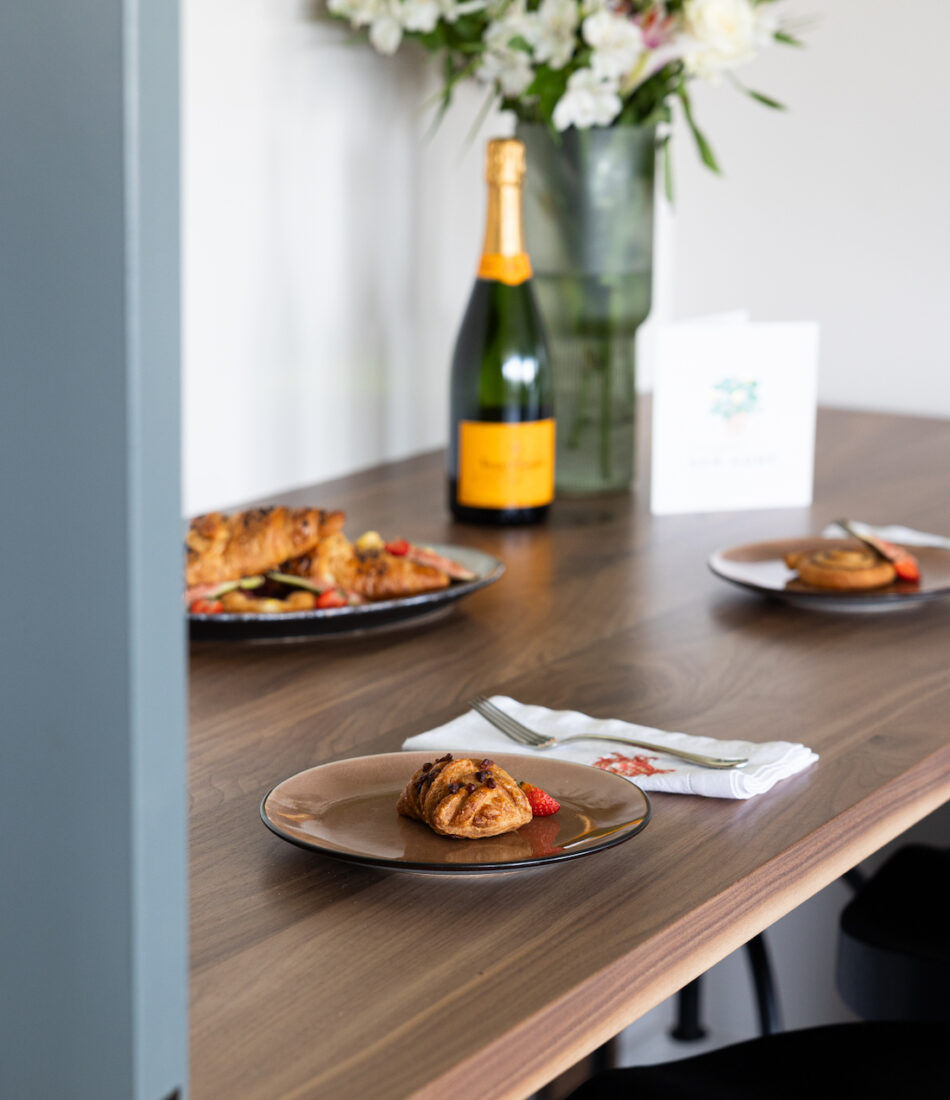 A wooden table displays plated pastries and strawberries, with a bottle of champagne and flowers in the background, suggesting a celebratory breakfast setting. A folded napkin and fork are nearby.
