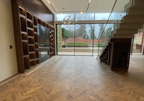A spacious, elegantly furnished room features wooden herringbone flooring. A large glass wall provides a view of an outdoor garden, enhanced by modern lighting and stylish shelving.