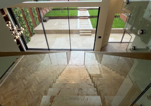 A marble staircase descends toward a spacious interior with large glass doors leading to a vibrant green garden, featuring a stone pathway and well-maintained landscaping.
