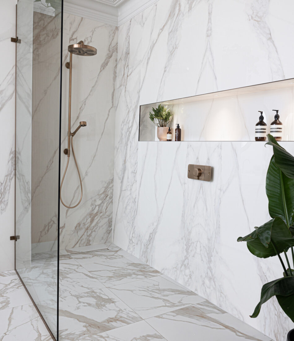 A marble-tiled bathroom features a glass shower with a bronze showerhead. In a recessed shelf, plants and decorative bottles sit, while a potted plant adds greenery to the space.