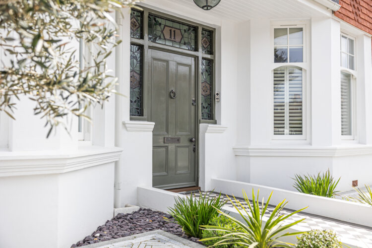 A green front door with a circular knocker is set in a white wall, flanked by decorative stained glass. Lush plants and decorative stones line the pathway leading to the entrance.
