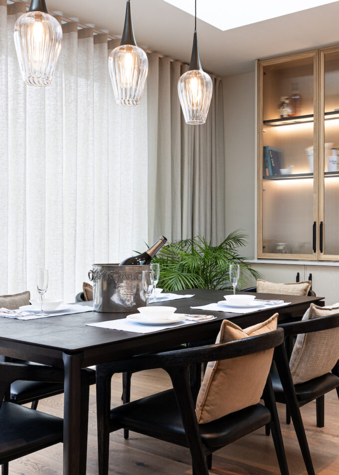 A black dining table is set with plates and glasses, holding an ice bucket with a bottle. Large windows with sheer curtains let in light, surrounded by modern decor and plants.
