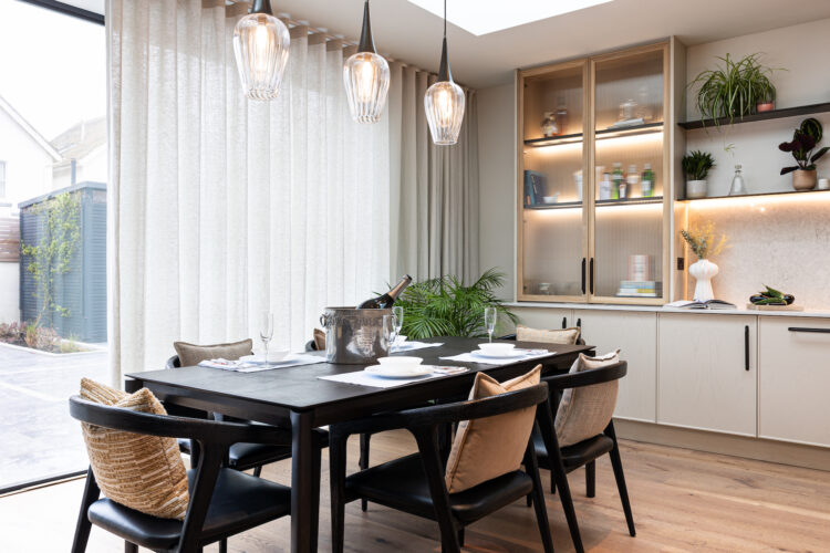 A black dining table is set with plates and glasses, holding an ice bucket with a bottle. Large windows with sheer curtains let in light, surrounded by modern decor and plants.