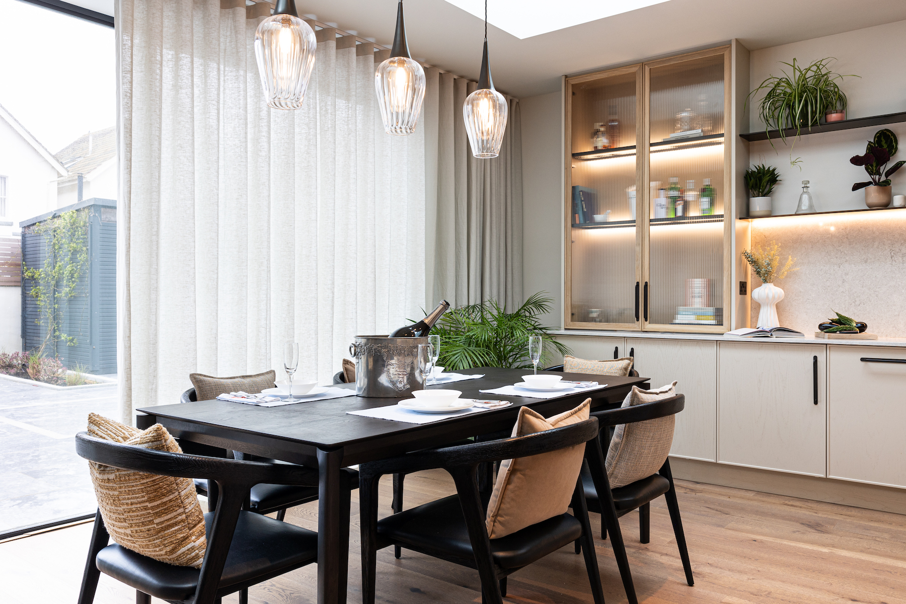 A black dining table is set with plates and glasses, holding an ice bucket with a bottle. Large windows with sheer curtains let in light, surrounded by modern decor and plants.