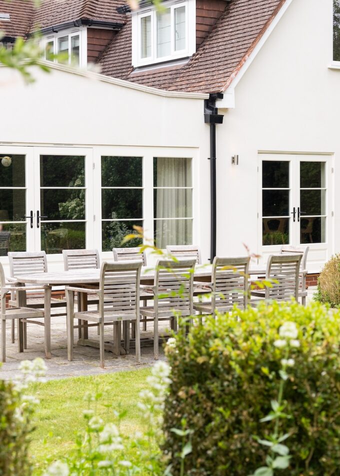 A wooden dining table with chairs is set outside a light-colored house. The scene is surrounded by manicured shrubs and colorful flowers, suggesting a serene garden setting.