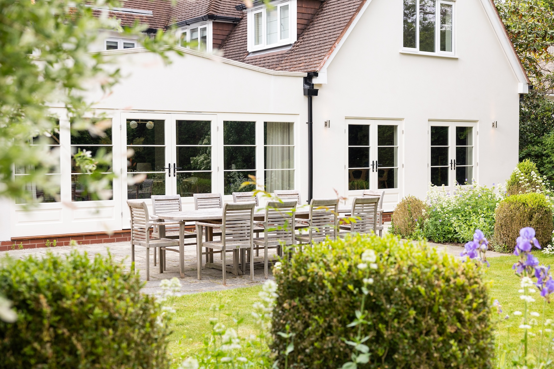 A wooden dining table with chairs is set outside a light-colored house. The scene is surrounded by manicured shrubs and colorful flowers, suggesting a serene garden setting.