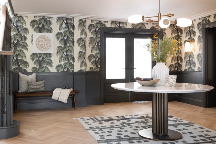 Art Deco Inspired Entrance Hall designed by Pfeiffer Design. Art deco inspired wallpaper, half height panelling and glazed French double doors in deep green. Bronze obscured glass wall lights and cream barnacle inspired artwork on walls. Central table with pendant light hanging down that match the wall lights with key bowl and decorative vase and candle. A timber bench for putting shoes on with cushion and throw scheme.