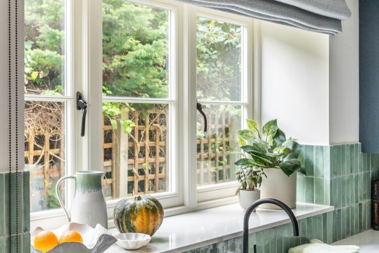 A kitchen window features plants and fruits on a marble countertop beneath a light gray roller shade. Natural greenery is visible outside, creating a fresh, inviting atmosphere.