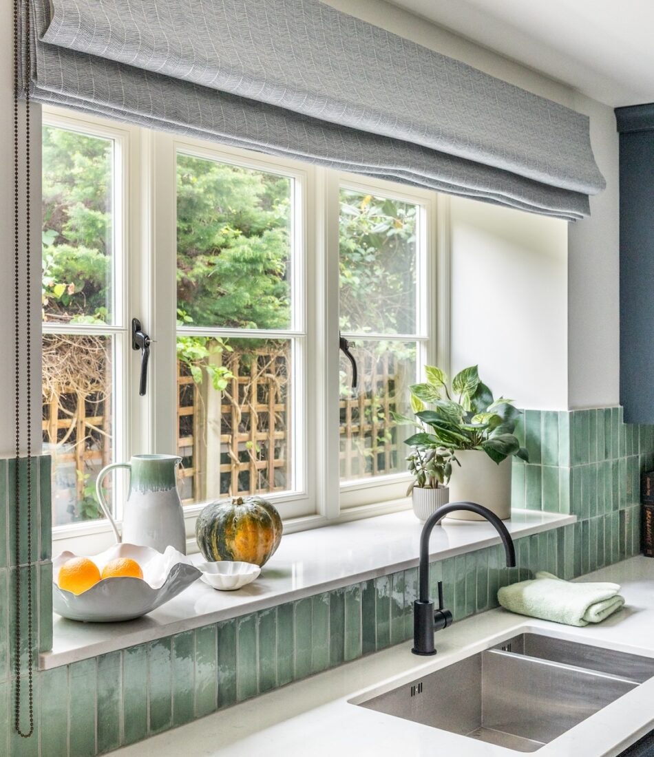 A kitchen window features plants and fruits on a marble countertop beneath a light gray roller shade. Natural greenery is visible outside, creating a fresh, inviting atmosphere.