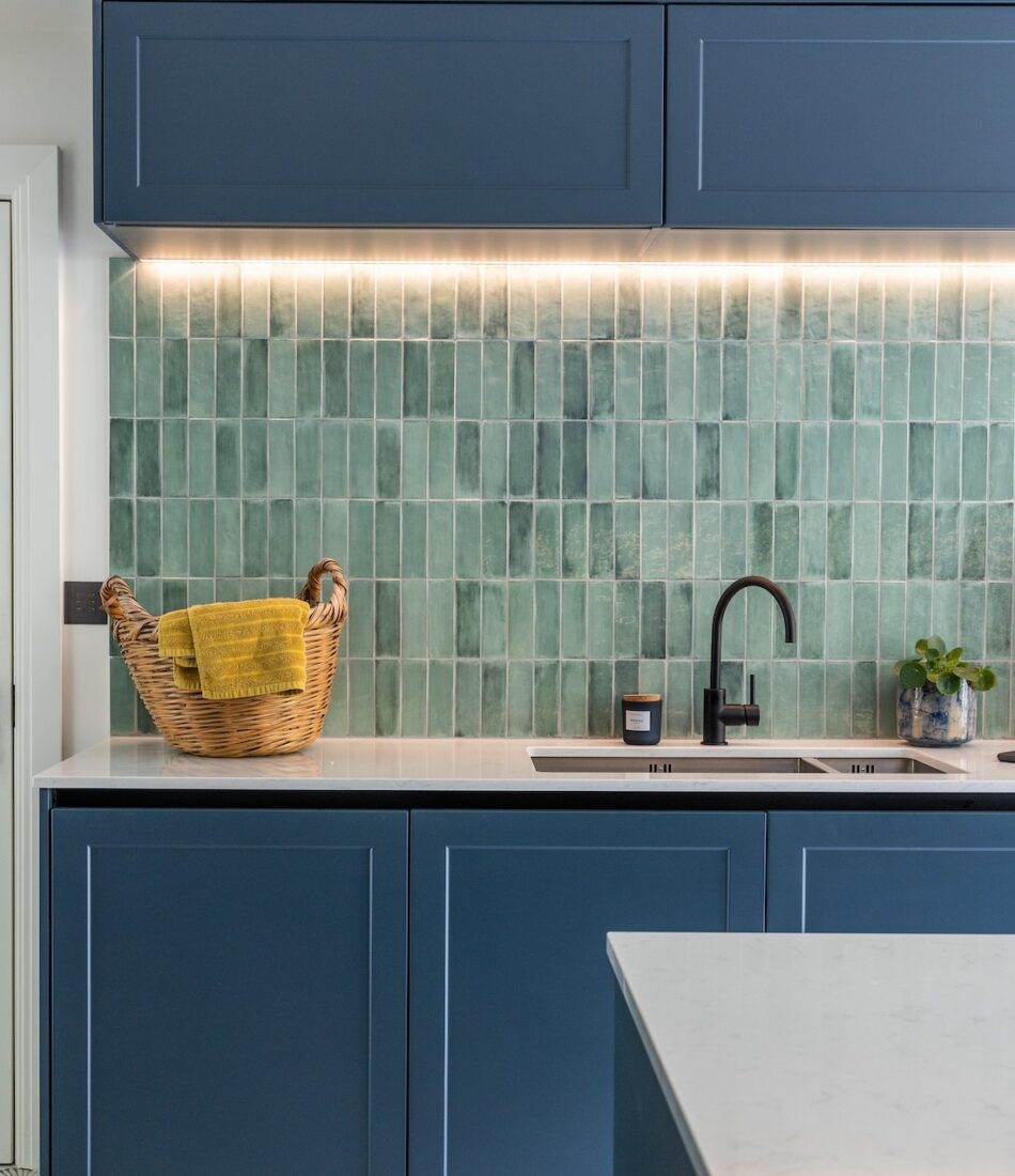 A wicker basket with a yellow towel sits on a countertop. Dual sinks and modern tap fixtures contrast against green tiled walls, illuminated by soft under-cabinet lighting in a sleek kitchen.