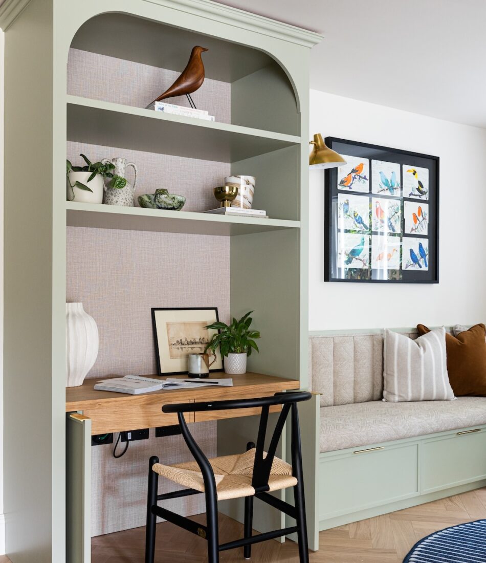 A green bookshelf with decorative items and plants surrounds a wooden desk and black chair, set against a light wall. A cozy, cushion-laden bench is nearby, enhancing comfort.