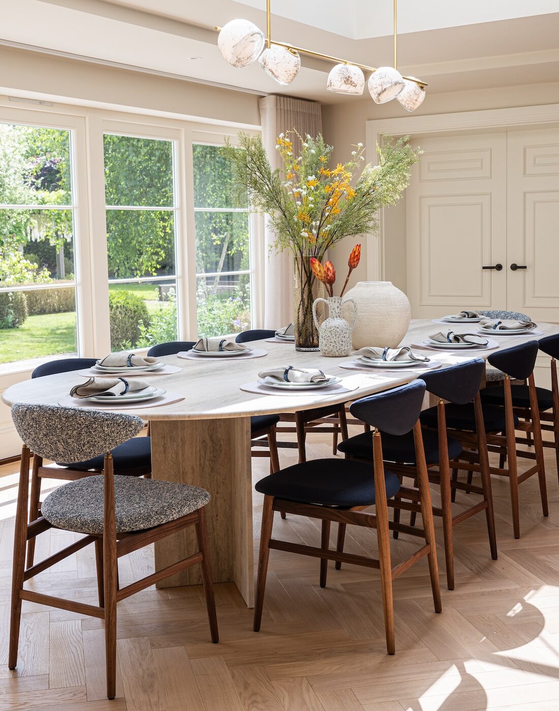 A round dining table is set with plates and cutlery, surrounded by high-backed chairs. A decorative vase with flowers is central, amidst a bright, sunlit room with large windows.