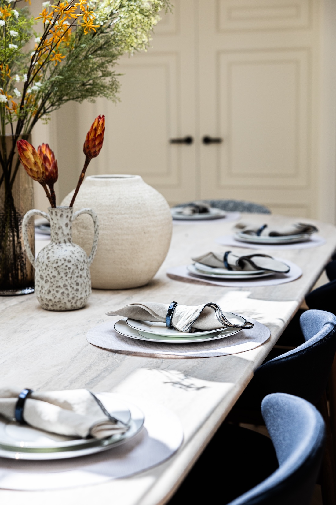 A dining table is set with plates and neatly folded napkins, surrounded by decorative vases filled with flowers. The background features elegant cabinetry, suggesting a modern, stylish kitchen environment.