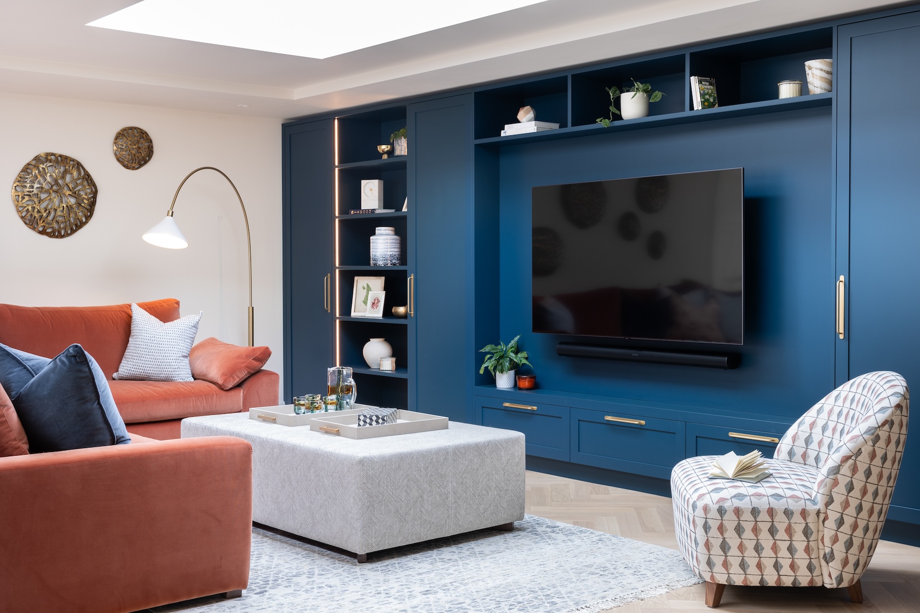 A cosy living room features a large blue entertainment unit with a television, an orange sofa, a patterned armchair, and a light-colored coffee table with a tray, surrounded by decorative elements.