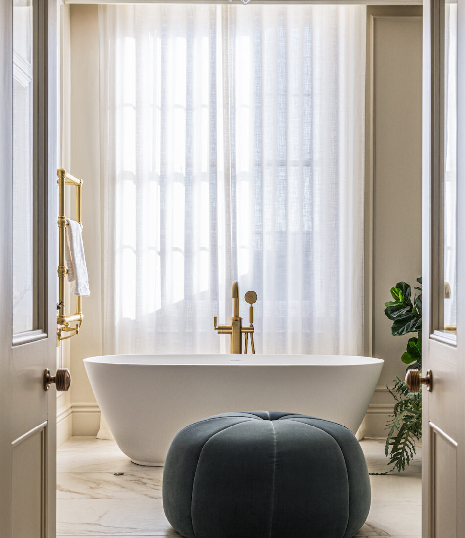 Principal En-suite with free standing bath, gold shower and towel rail, navy blue stool, long linen drapes with light shining through window