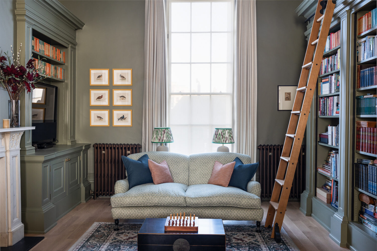 Library design, green walls with green textured sofa and lamp shades with contrasting cushions. Tall rolling ladder for access to book shelves