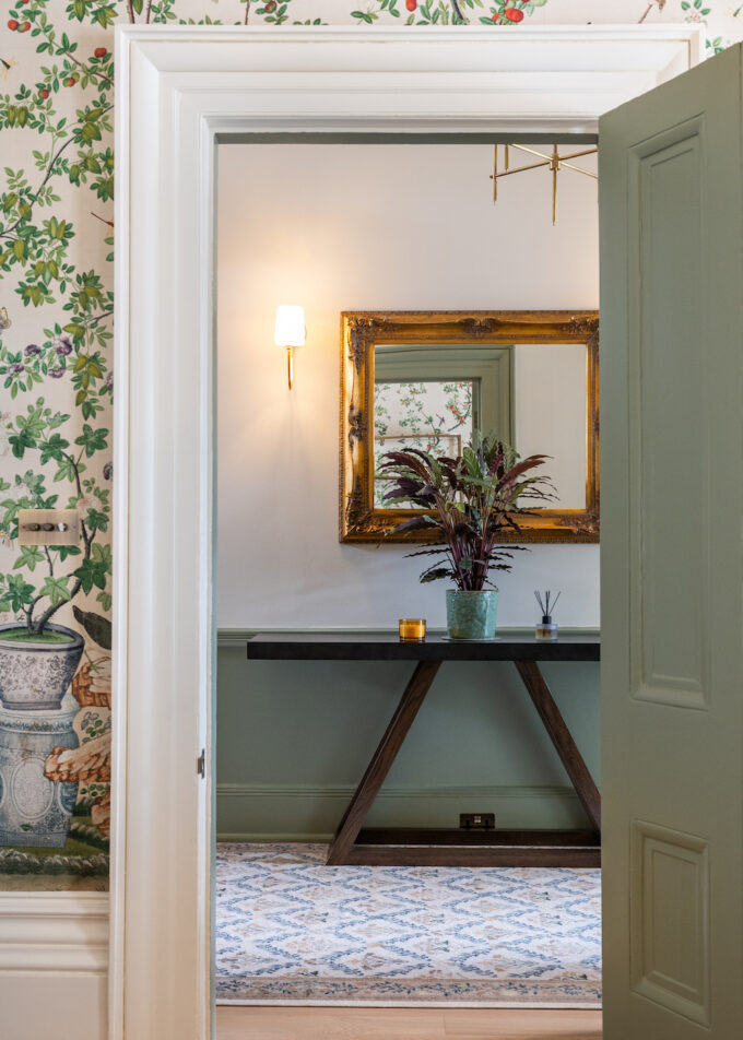 View through interior doorway into the corridor - looking at wooden console table and gold framed mirror with wall lights and plants