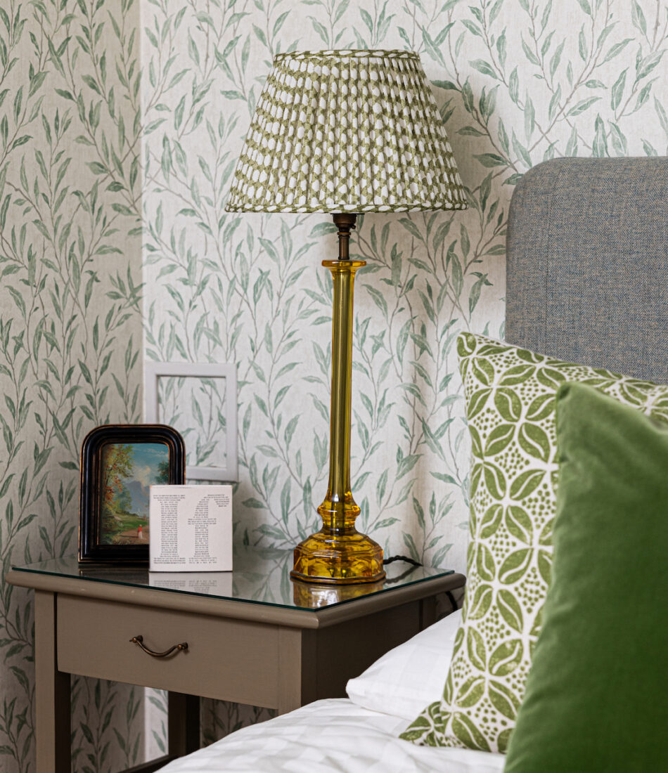 Green pleated lamp shade on bedside table blending with green floral wallpaper behind and green bedding