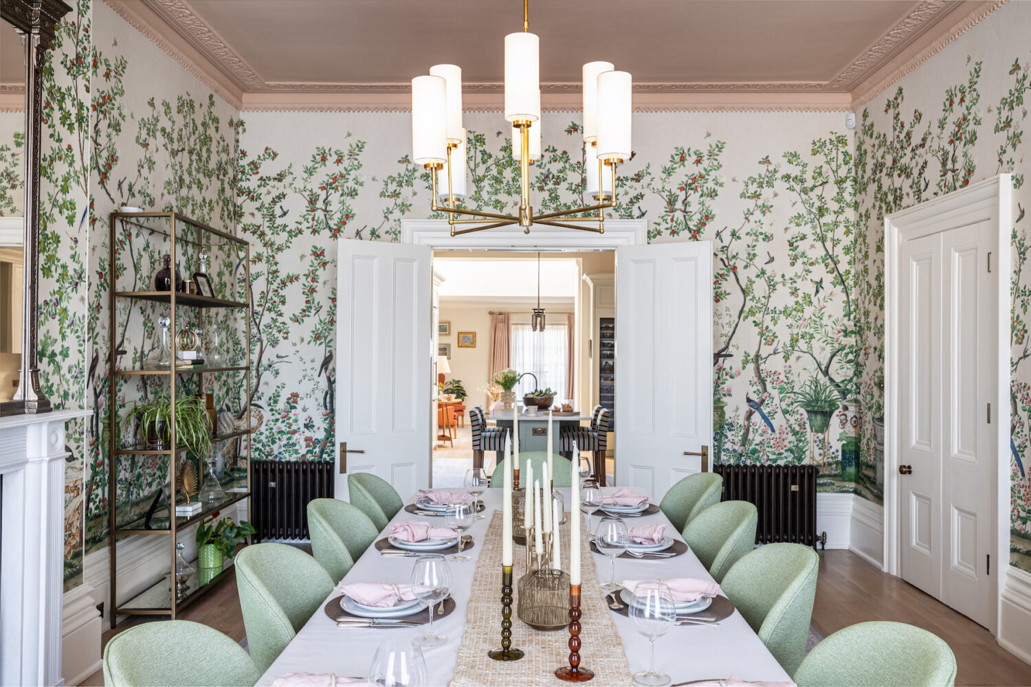 Grand Regenecy townhouse dining room with pink ceiling and cornice, bold patterned tree wallpaper and a set dining table with green chairs, candles, crockery