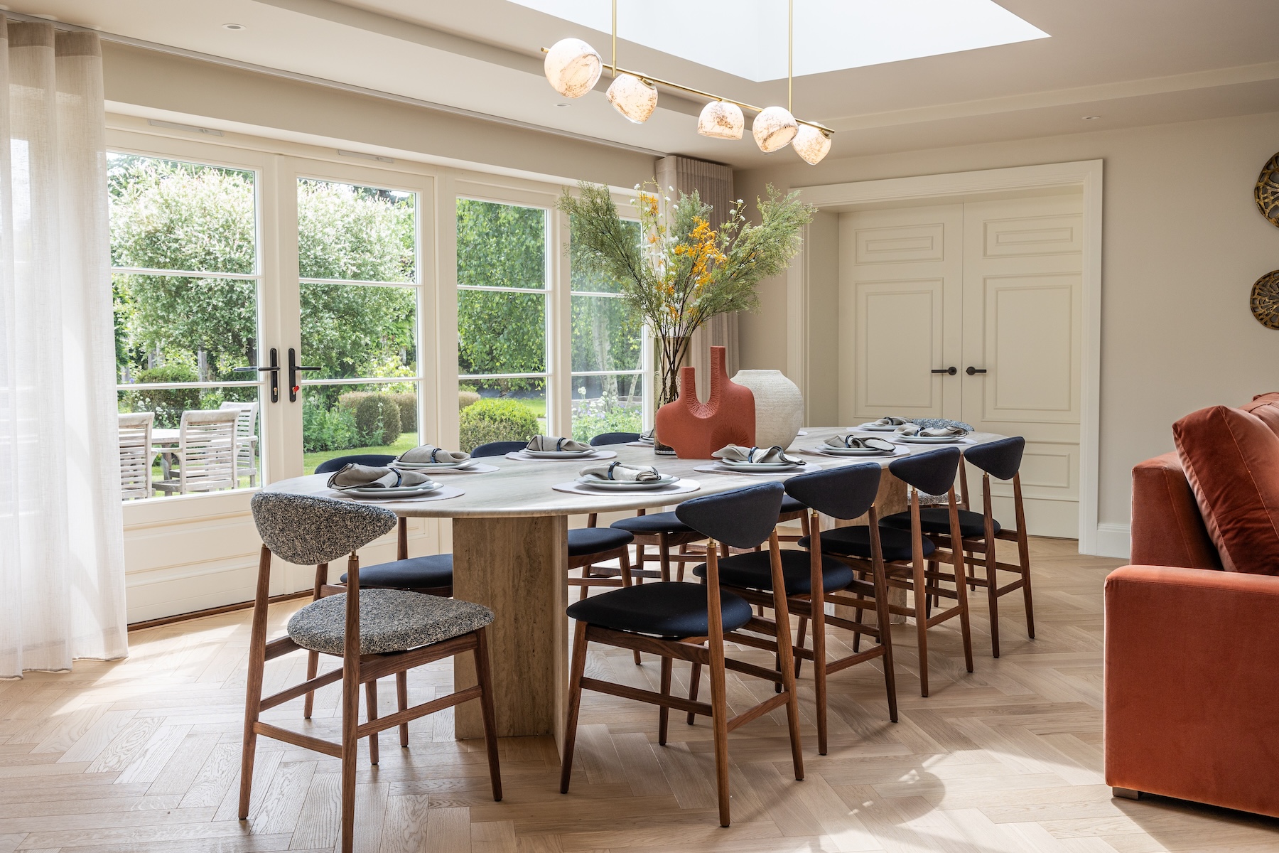 A round dining table is set with plates and cutlery, surrounded by high-backed chairs. A decorative vase with flowers is central, amidst a bright, sunlit room with large windows.