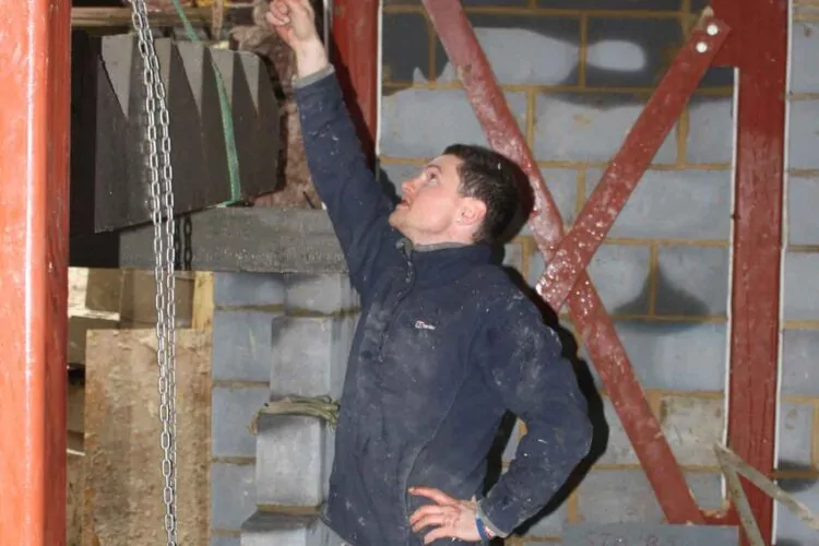 A man points upward while standing in a construction site, surrounded by unfinished walls with various building materials and support structures, indicating focus and communication regarding the task.
