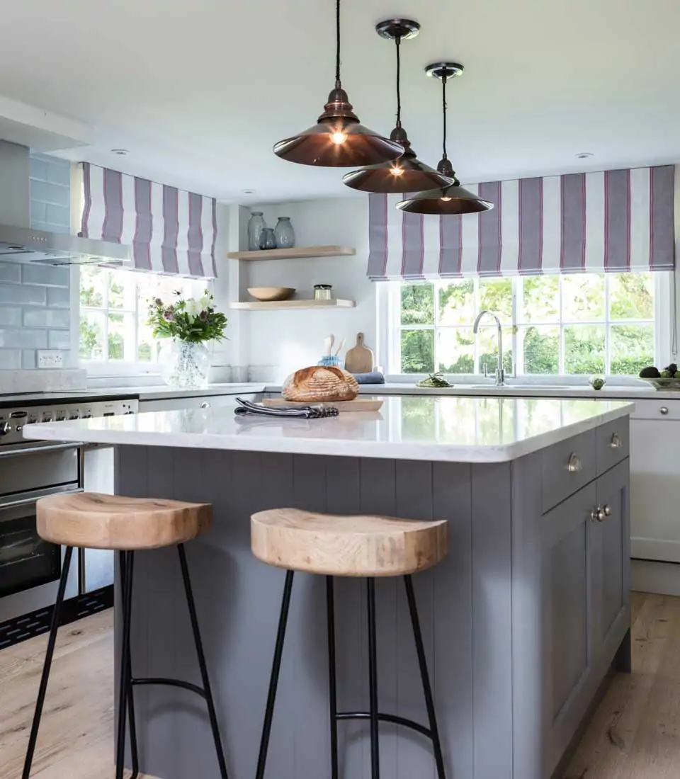 A modern kitchen features a central gray island with a loaf of bread, two wooden bar stools, and three pendant lights hanging above. Natural light streams through window blinds.