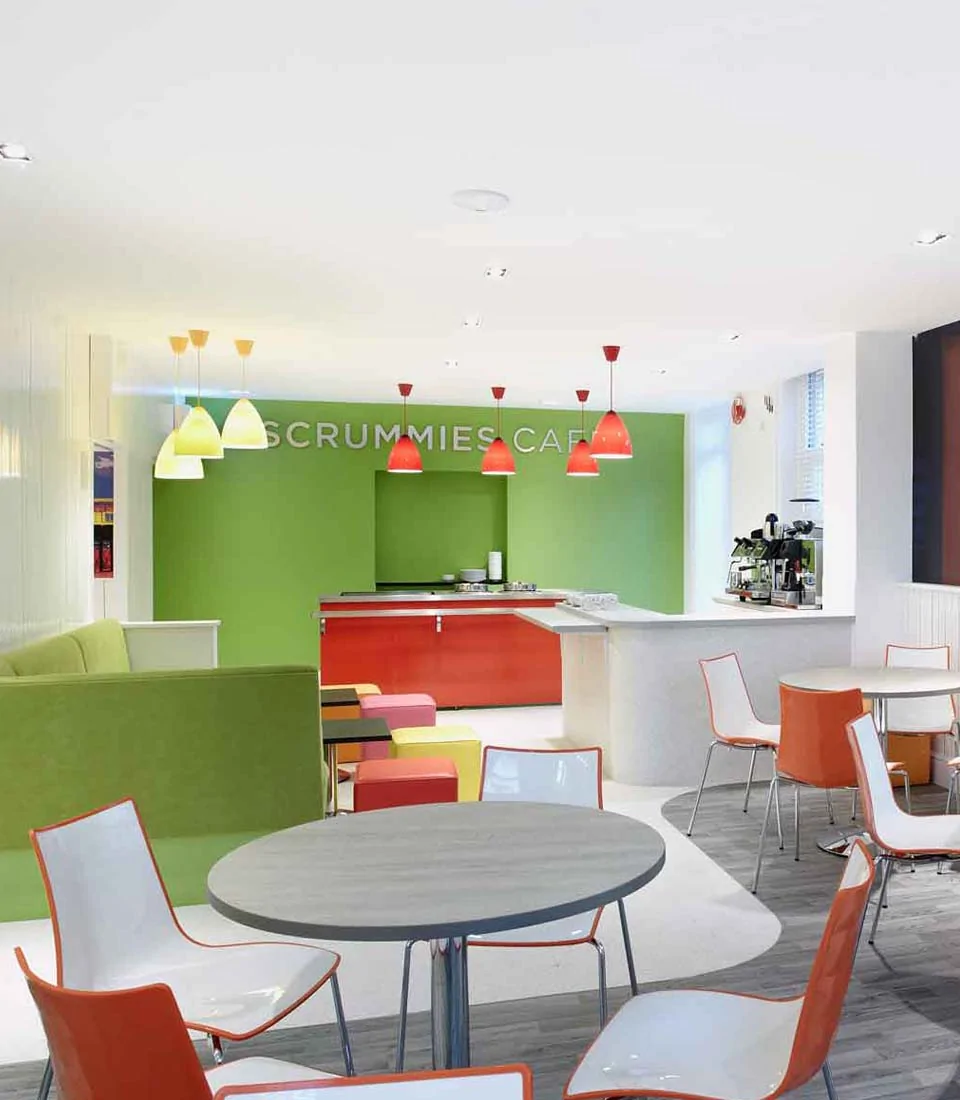 A bright café features a green accent wall and colorful pendant lights. Tables and chairs are arranged for seating. An open counter with kitchen equipment is in the background, labeled "SCRUMMIES CAFÉ."