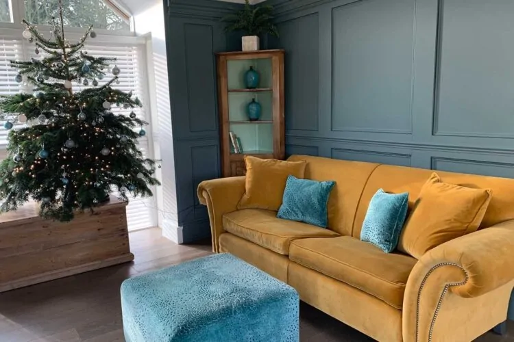 A yellow velvet sofa with blue cushions sits in a cozy room. A small teal ottoman rests in front, while a decorated Christmas tree is positioned nearby, surrounded by natural light.