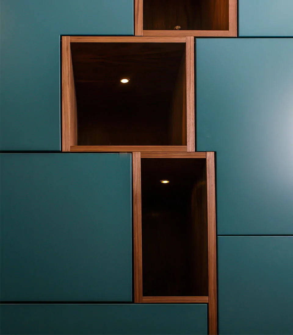 A series of rectangular wooden shelves, inlaid within a teal wall, feature warm lighting inside some spaces, creating an elegant contrast in the modern interior design.